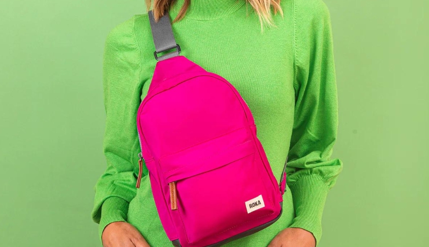 Lifestyle image of Roka Willesden Candy Pink Rucksack with single strap. With green background and lady wearing green jumper and rucksack forward facing.